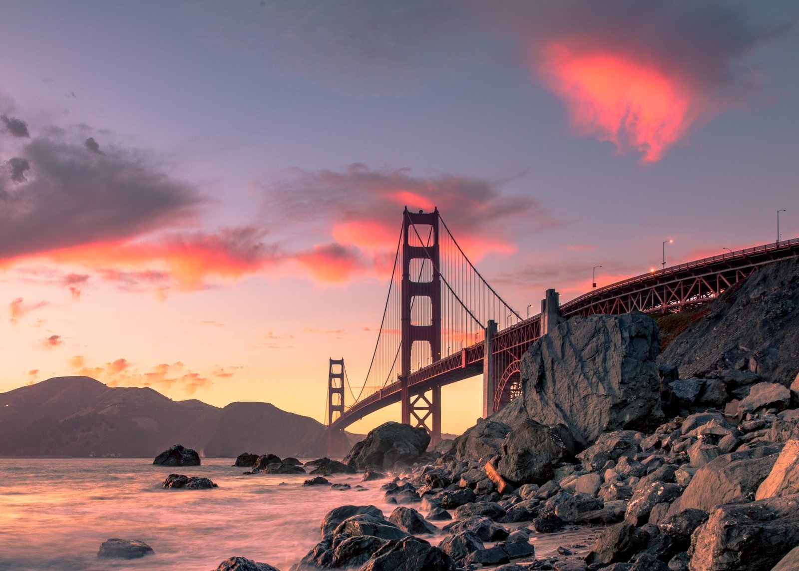 A wide shot of Golden Gate Bridge on the body of water near rock formations during sunset in San Francisco, California